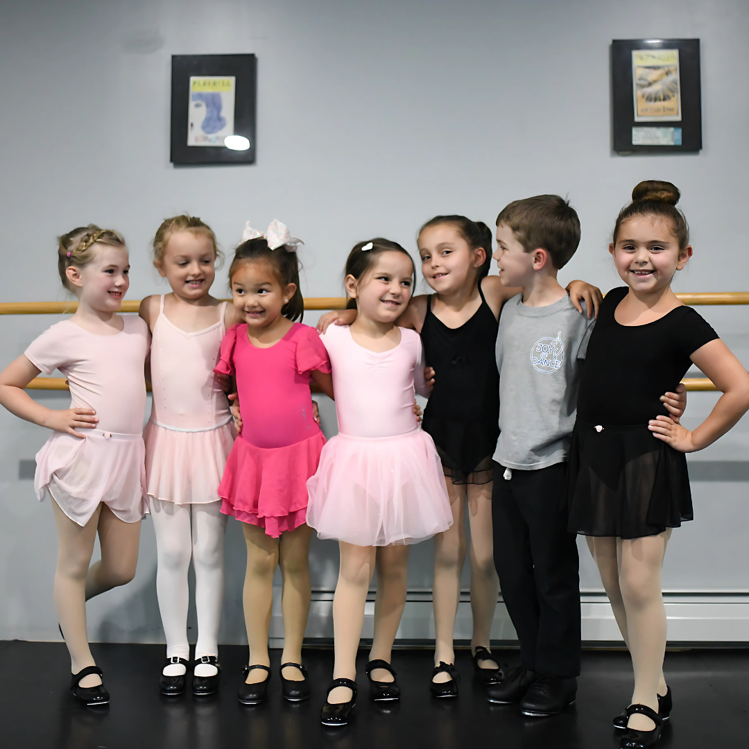 Group of adult ballet dancers posing playfully in a dance studio with wooden barres, blue walls, and a screen in the background.