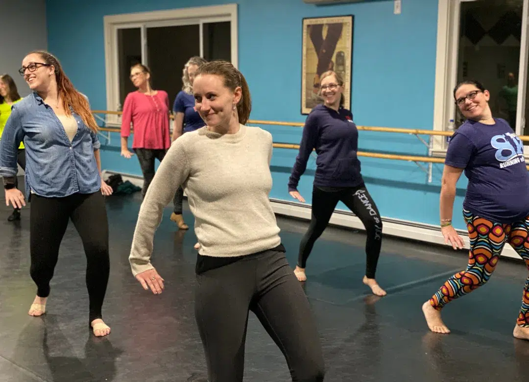 Group of adult ballet dancers posing playfully in a dance studio with wooden barres, blue walls, and a screen in the background.