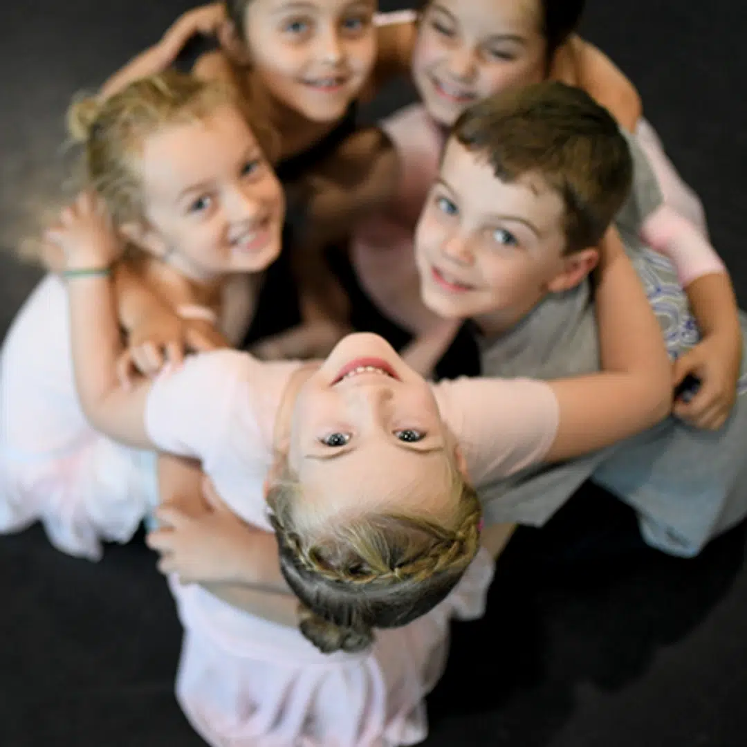 Group of adult ballet dancers posing playfully in a dance studio with wooden barres, blue walls, and a screen in the background.