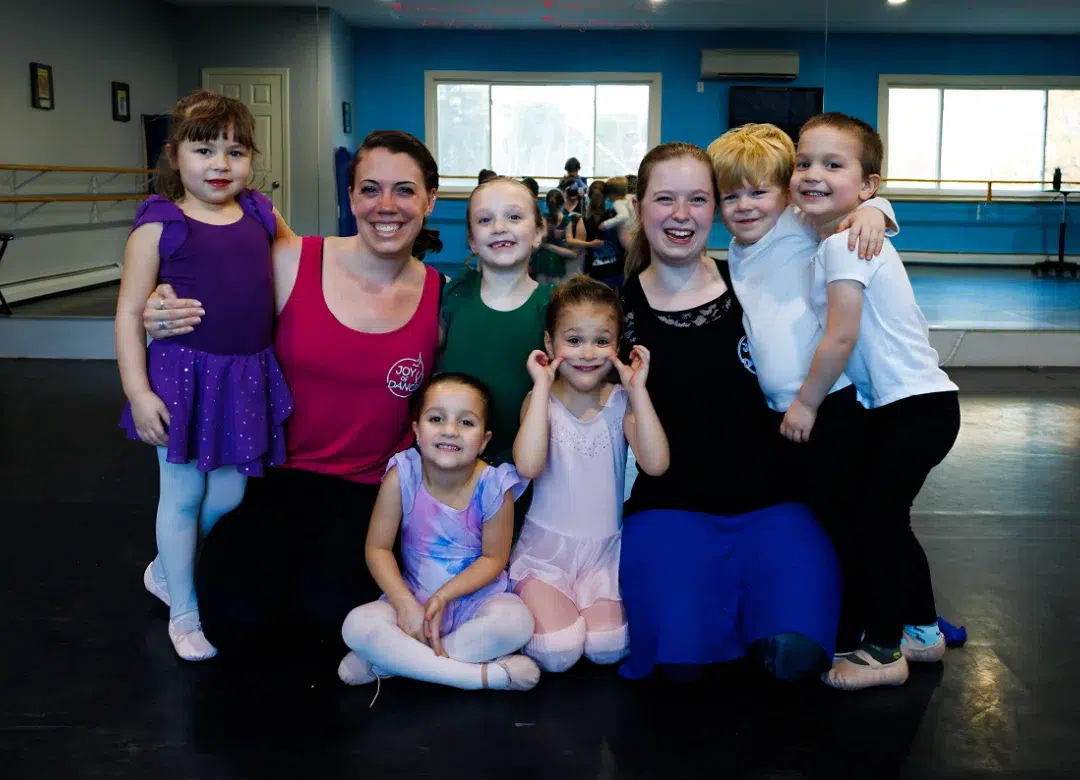Group of adult ballet dancers posing playfully in a dance studio with wooden barres, blue walls, and a screen in the background.